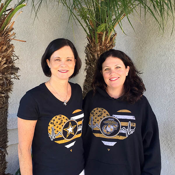 an army mom and marine mom wearing black shirts with heart designs and symbols, standing against a light-colored wall with plants.
