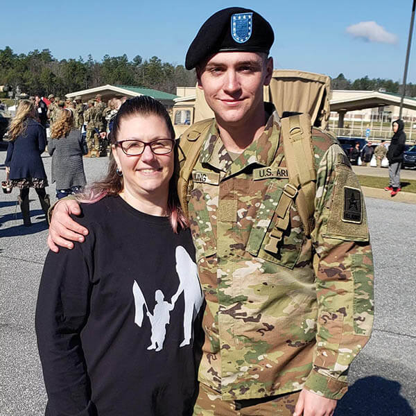 Army mom hugging her Soldier in uniform outside a building with people in the background.