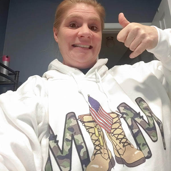 army mom wearing a white hoodie with military-themed graphics and text, giving a thumbs-up.